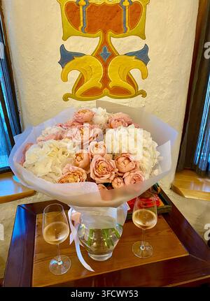 Elegant bouquet of roses and hydrangeas stands beside two wine glasses indoors. Stock Photo