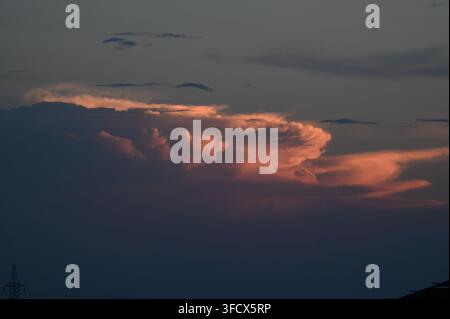 View of dramatic sunset clouds with pink and orange hues illuminated by the setting sun against a dark sky, with a power pylon silhouette in Jahanian, Stock Photo