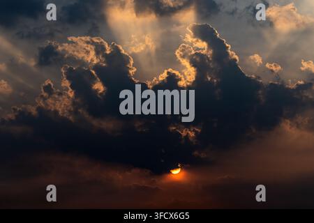 View of the sun setting behind dramatic, dark storm clouds with golden light rays piercing through the sky in Jahanian, Punjab, Pakistan. Stock Photo