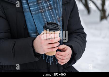 Close up of man holding takeaway coffee cup with both hands outdoors in snowy winter park. Hot drink, cold weather and winter lifestyle concept. Stock Photo