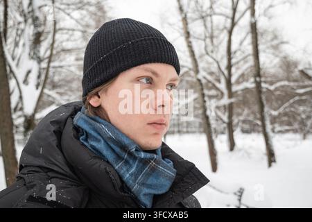 Close up portrait of young man wearing black hat and blue scarf in snowy park. Natural winter lifestyle portrait in cold weather with copy space. Stock Photo