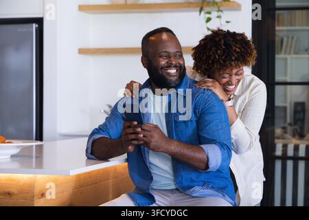 African American couple laughing, man holding smartphone while sitting on stool at kitchen island Stock Photo