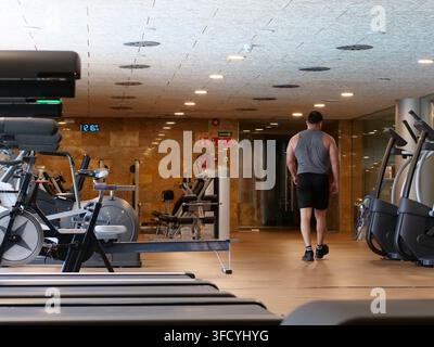 Rear view of an athletic male figure walking away through an empty modern gym aisle. Surrounded by professional cardio equipment, treadmills, and resi Stock Photo