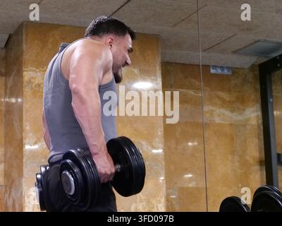 Side view of a strong muscular man performing dumbbell shrugs with high effort and tension in a modern gym studio. Focused on shoulder and trapezius m Stock Photo