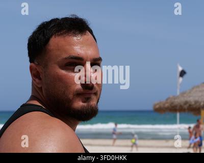 Portrait of a muscular man in black tank top standing on a sunny beach, turning to look directly at the camera. Background features turquoise ocean wa Stock Photo