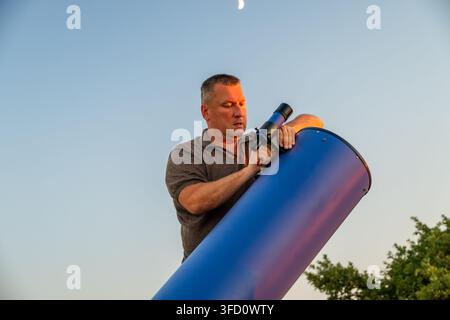 a man adjusting a large blue telescope outdoors during twilight with a crescent moon visible in the sky. Stock Photo