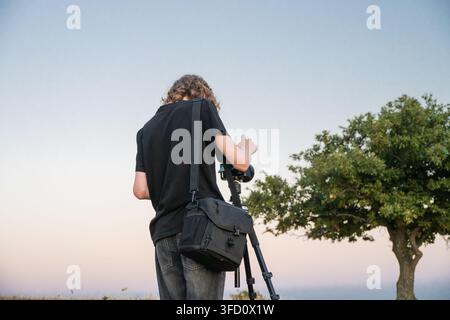 a person with a camera bag over their shoulder shooting photographs with a camera mounted on a tripod outdoors. Stock Photo