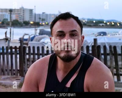 Portrait shot of a bearded handsome athletic man looking directly at camera. Taken outdoors near coastal harbor marina with blurred boats and resort b Stock Photo
