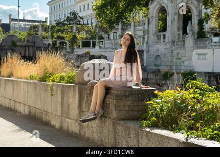 Full length portrait of smiling woman sitting on stone wall in sunny park. Stock Photo