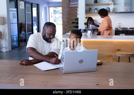 African American family working at wood table in open plan home, man writing notebook. Collaboration, learning, laptop, tablet, kitchen, island, dayli Stock Photo