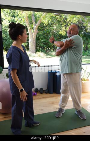 Diverse pair in clinic on mat, therapist in scrubs guiding client doing shoulder stretch by window. Therapy, rehab, fitness, mobility, oneonone, dayli Stock Photo