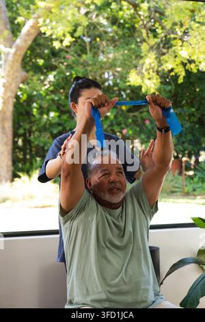 Senior man seated raising blue exercise band by window while Asian caregiver in scrubs guiding arms Stock Photo