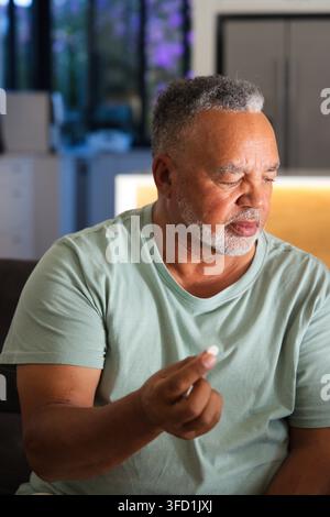 Senior African American man holding small white pill, sitting on dark couch near kitchen cabinets Stock Photo