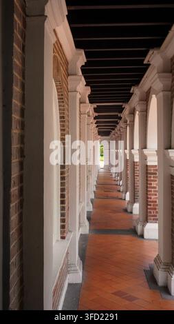 Colonial Building Corridor with White Pillars and Brick Walls. Vertical perspective view of a long empty colonial-style corridor with white columns, r Stock Photo