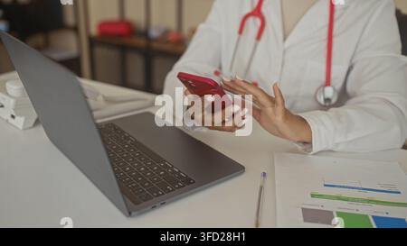 Woman doctor taps red smartphone with hands and stethoscope beside laptop on desk inside building; focus. Stock Photo