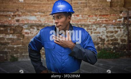 Man construction worker, african american, in blue hardhat and blue coveralls, places hand on chest in front of brick building, serious expression and Stock Photo
