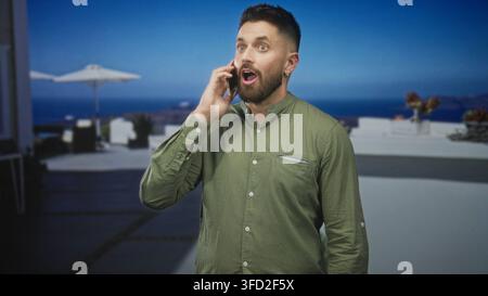 Bearded hispanic man tapping smartphone screen with thumb while smiling, holding phone in left hand on building terrace overlooking sea; joy connectio Stock Photo