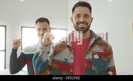 Two men showing i voted sticker and raised fists, one hand clenched near chest in polling building; civic pride. Stock Photo