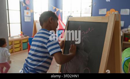 African american boy erases a chalkboard with hand while drawing a house on an easel in a daycare building filled with toys and bunting; playful focus Stock Photo