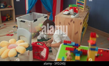 Boy kneeling and opening red toy case with hands beside blocks and plush, focused on the handle in a playroom inside a building; quiet curiosity. Stock Photo