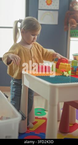 Girl stacking toy blocks on a small table in a building playroom, wearing yellow sweater and jeans; concentration creativity learning. Stock Photo