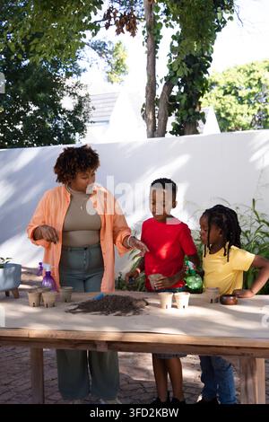 African American woman guiding two children planting soil on paved patio with trowel and cups Stock Photo