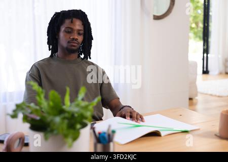 Adult man wearing olive T-shirt and bracelet sitting at wooden table at home writing in notebook Stock Photo