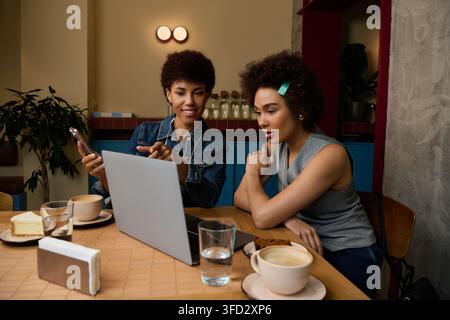 Two young African American women laugh and share ideas over coffee while using a laptop. Stock Photo