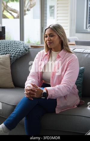 African American woman sitting on grey sofa in living room, wearing pink shirt, smartwatch, throw. Interior, home, natural, daylight, neutral, pastel, Stock Photo