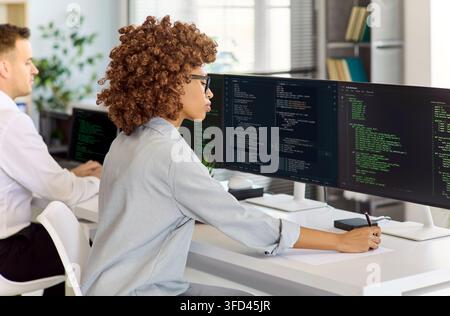 Female software developer coding at multi monitor workstation in modern tech office Stock Photo