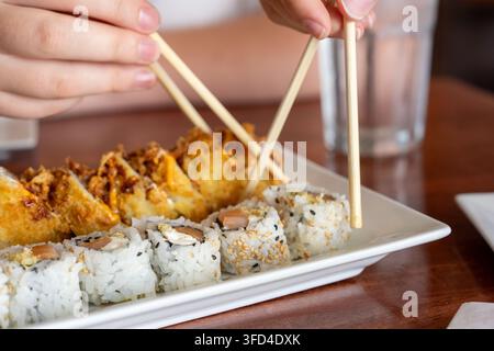 Close-up of two people using wooden chopsticks to pick up sushi from a shared plate in a Japanese restaurant. Assorted salmon rolls and crispy fried s Stock Photo