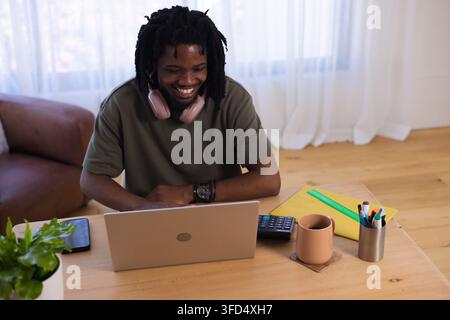 African American man smiling, leaning, using silver laptop at home table with pink headphones, mug Stock Photo