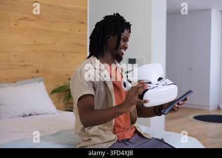 Male sitting on bed edge inspecting white VR headset and tablet in modern bedroom Stock Photo