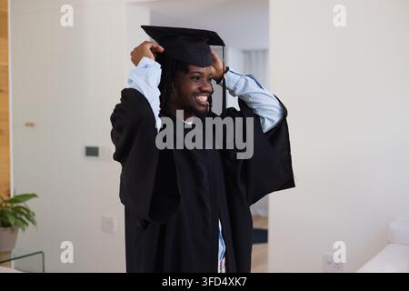 Man standing and adjusting mortarboard while smiling in black graduation gown in modern living room Stock Photo