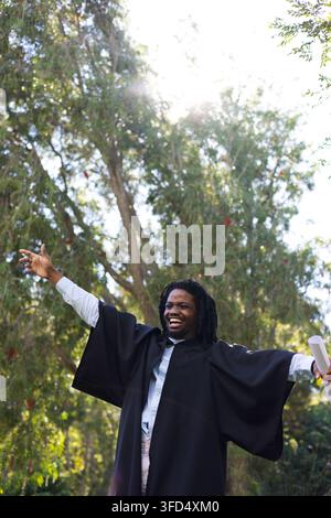 Adult African man standing outdoors among tall trees wearing black academic gown holding diploma Stock Photo
