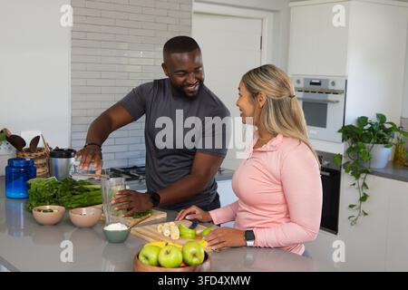 Couple preparing smoothie at island, man pouring blender jar, woman in pink top slicing apples Stock Photo
