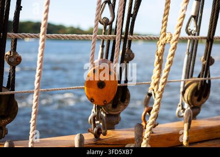 Wooden pulley block, belaying pins, and shroud ropes on the wooden deck rail of a sailboat with blurred blue sea water in background. Stock Photo