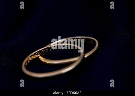 Two thin gold wedding rings with engraved text resting on a black background, softly lit.​ Stock Photo