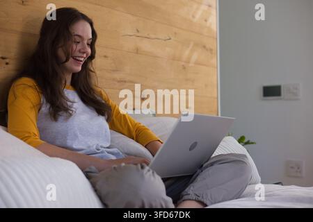 Adult female sitting on bed using silver laptop, smiling with pillows, plant, thermostat Stock Photo