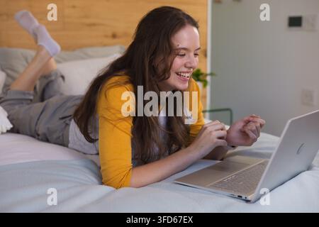 Woman wearing mustard-yellow top and gray overalls lying on stomach in bedroom using silver laptop Stock Photo