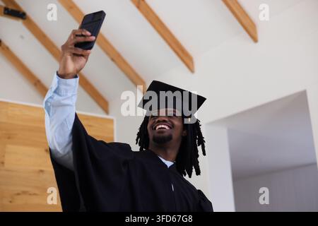 African American man smiling raising phone taking selfie in room with beams mortarboard tassel gown Stock Photo