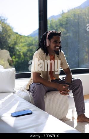 African American man sitting on ottoman at home, wearing orange tee, pink headset, phone on couch Stock Photo