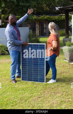 African American homeowner and solar technician holding solar panel, gesturing on grassy lawn Stock Photo