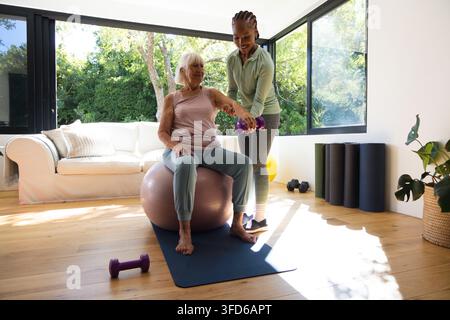 Diverse female trainer supporting senior woman lifting purple dumbbell on stability ball at home Stock Photo
