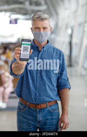 A man wearing a face mask holds up his mobile phone displaying a digital health pass or vaccination record with a QR code Stock Photo