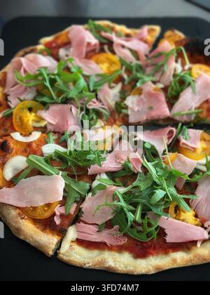 Homemade pizza topped with sliced ham, yellow tomatoes, and fresh arugula. Freshly baked homemade pizza sliced on a dark surface, featuring a golden c Stock Photo