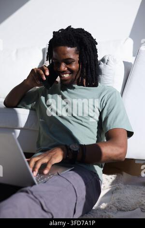 Man reclining on sofa at home in sage tee, talking on smartphone and typing on laptop Stock Photo