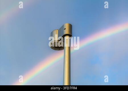Low angle view of a modern outdoor security camera on a metal pole beneath a bright rainbow and clear blue sky. Stock Photo