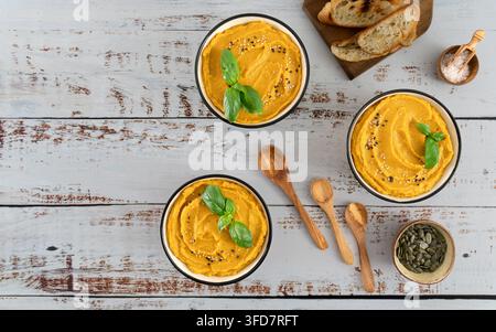 Overhead view of creamy pumpkin puree garnished with basil sesame seeds and pumpkin seeds served with toasted bread crisps on a rustic white wooden ta Stock Photo