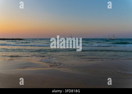 A bright orange sun sets low over the sea, glowing behind the silhouette of a rocky breakwater and gentle waves. Stock Photo